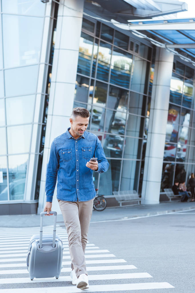 Красивый взрослый мужчина с багажом, пересекающий пешеход, заглядывающий в смартфон на заднем плане аэропорта
 - Фото, изображение