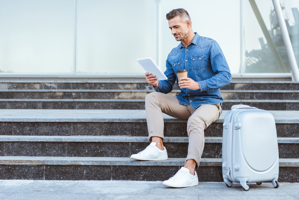schöner erwachsener Mann mit einem Coffee to go auf der Treppe sitzend mit Gepäck und digitalem Tablet - Foto, Bild