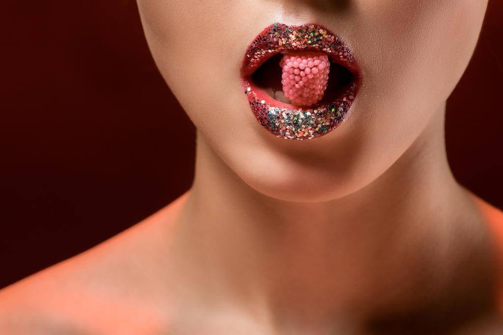 молода жінка з блискучими губами тримає рожеві цукерки в роті на бордовому фоні
 - Фото, зображення