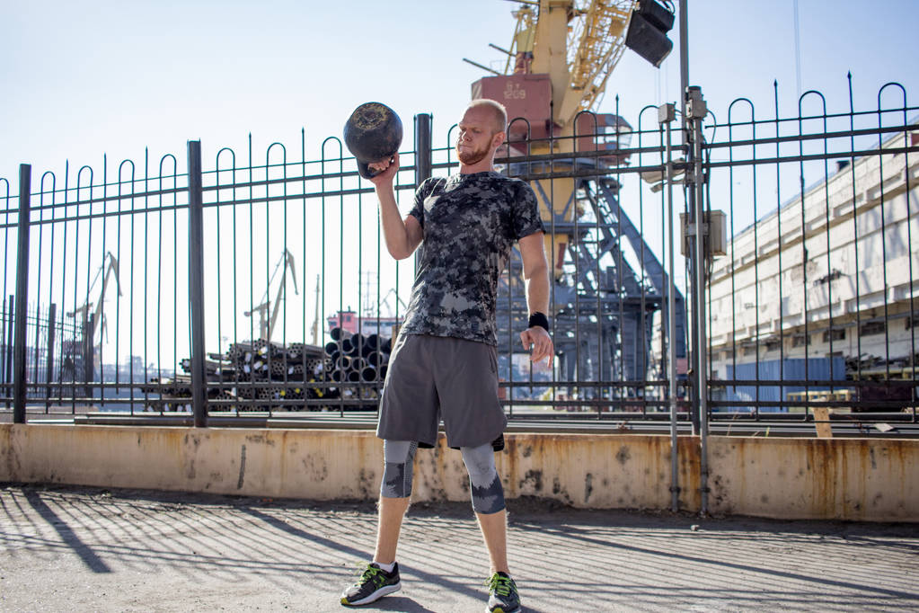 Young bearded male athlete training in industrial zone in sunny day, kettlebells exercises outdoors, urban background - Photo, Image