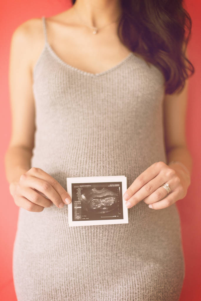 lachende zwangere vrouw weergegeven: echografie foto van haar ongeboren baby geïsoleerd op rode achtergrond - Foto, afbeelding