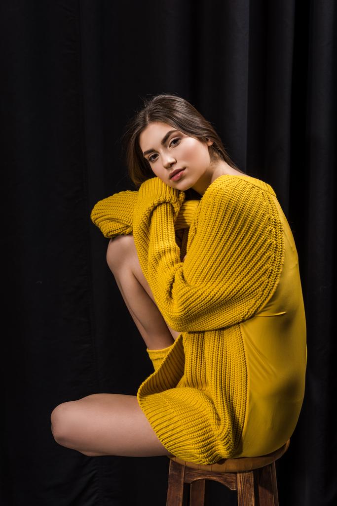 side view of pensive woman in yellow woolen sweater on wooden bar stool on black backdrop - Photo, Image