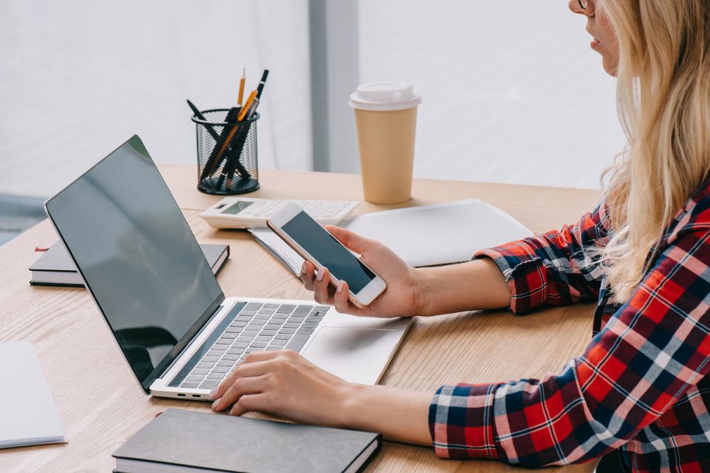Teilbild einer Geschäftsfrau, die Smartphone und Laptop gleichzeitig am Arbeitsplatz mit Papieren im Büro benutzt - Foto, Bild