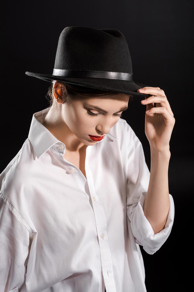 stylisches junges Model mit roten Lippen in weißem Hemd und schwarzem Hut posiert isoliert auf schwarz - Foto, Bild