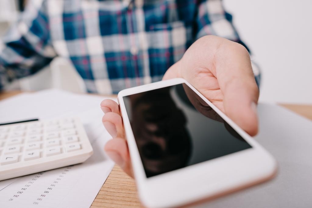 Schnappschuss von Geschäftsmann zeigt Smartphone mit leerem Bildschirm am Arbeitsplatz mit Papieren und Taschenrechner - Foto, Bild