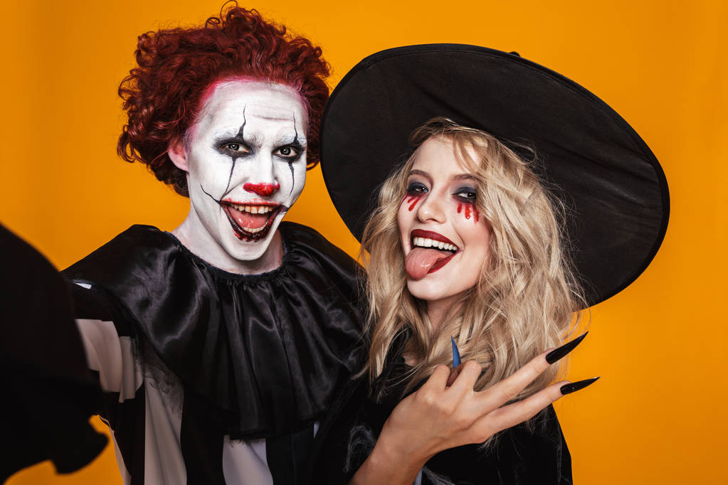 Foto van vrolijke heks vrouw en clown man dragen zwart kostuum en halloween make-up nemen selfie geïsoleerd op gele achtergrond - Foto, afbeelding