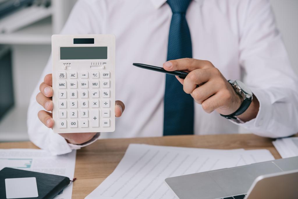 частичный взгляд бизнесмена, указывающего на калькулятор в руке на рабочем месте, концепция бухгалтерского учета
 - Фото, изображение