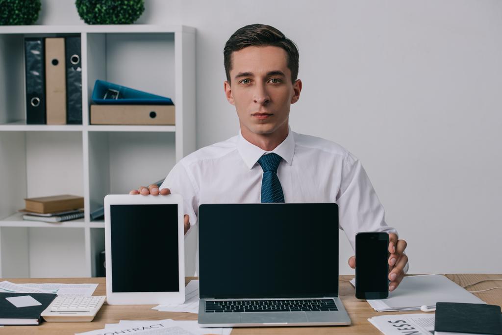 portrait of businessman showing laptop, tablet and smartphone with blank screens at workplace in office - Photo, Image