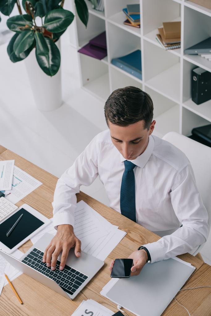 високий кут зору бізнесмена за допомогою смартфона і ноутбука на робочому місці з документами в офісі
 - Фото, зображення