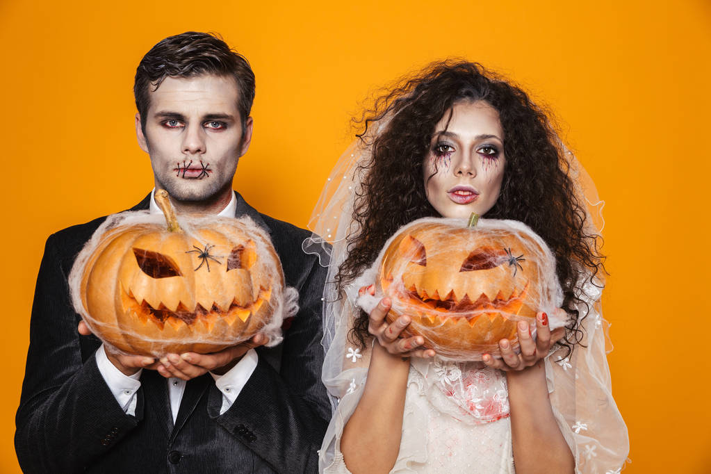 Photo of scary zombie couple bridegroom and bride wearing outfit and halloween makeup holding carved pumpkin isolated over yellow background - Photo, Image