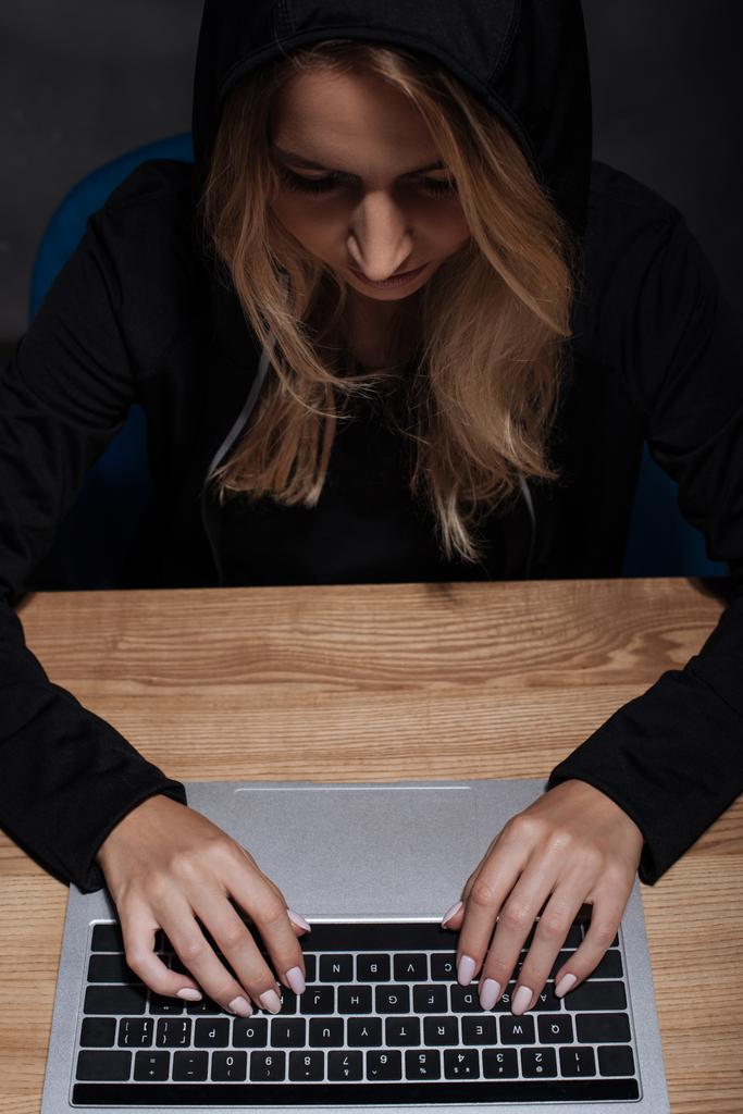 hacker feminino com capuz preto usando laptop em mesa de madeira
 - Foto, Imagem