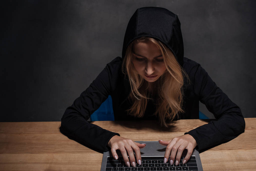 hacker femelle concentrée en sweat à capuche noir à l'aide d'un ordinateur portable sur table en bois
 - Photo, image