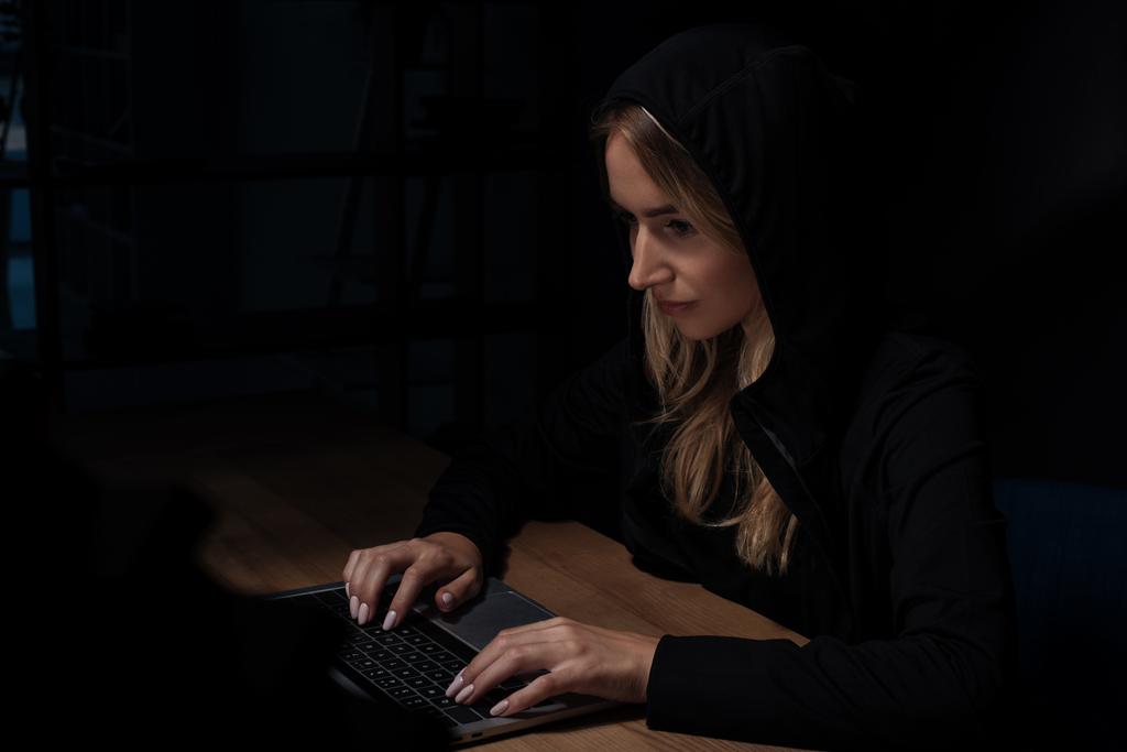 vista lateral de la mujer en sudadera con capucha negra usando el ordenador portátil, concepto de seguridad cibernética
 - Foto, imagen
