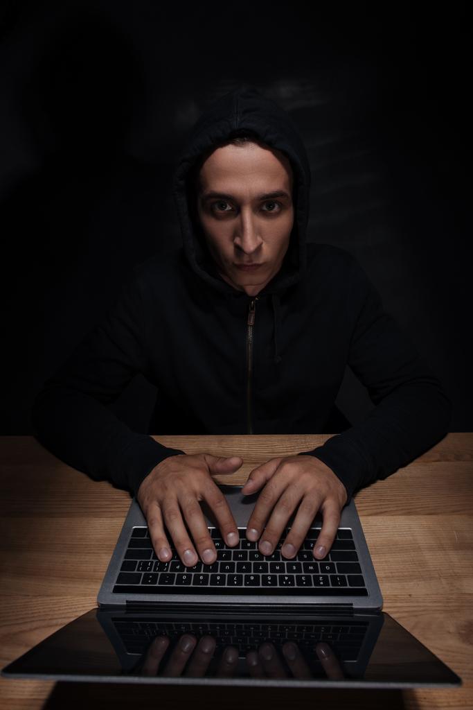 retrato de hacker en sudadera con capucha negro mirando a la cámara mientras se utiliza el ordenador portátil, concepto de seguridad del cuber
 - Foto, Imagen
