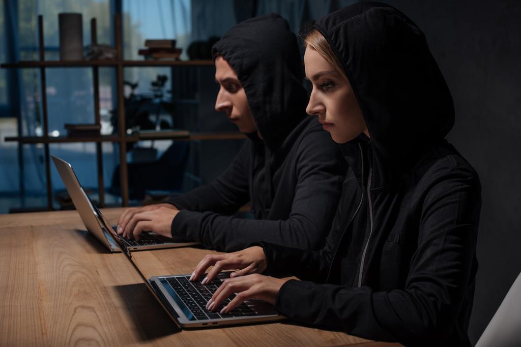 Seitenansicht von Hackern in schwarzen Kapuzenpullovern mit Laptops an hölzerner Tischplatte, Cyber-Sicherheitskonzept - Foto, Bild