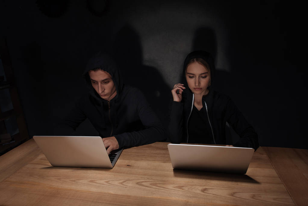 hackers in black hoodies using laptops at wooden tabletop in dark room, cyber security concept - Photo, Image