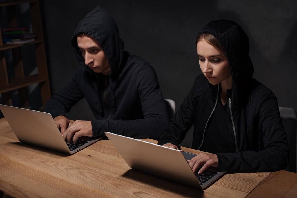hackers in zwarte hoodies met behulp van laptops op houten tafelblad, cyber veiligheidsconcept - Foto, afbeelding