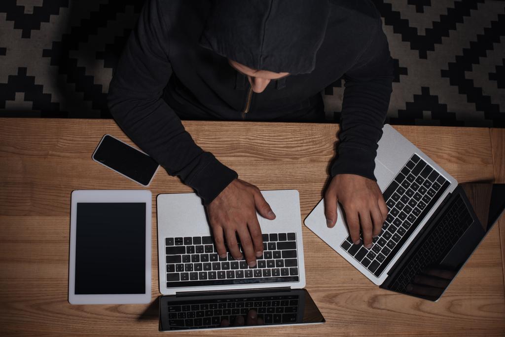 vista aérea de hacker en sudadera con capucha negra usando computadoras portátiles en la mesa con tableta y teléfono inteligente, concepto de seguridad cibernética
 - Foto, Imagen
