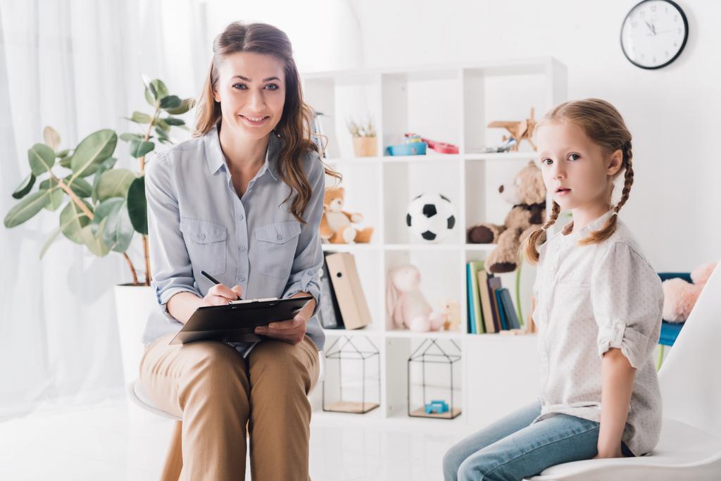 smiling psychologist with clipboard sitting in office with little child and looking at camera - Photo, Image
