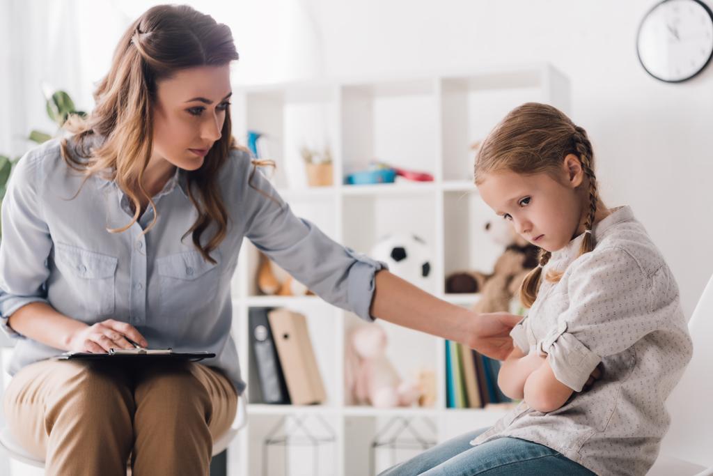 adult psychologist with clipboard talking to sad little child in office - Photo, Image