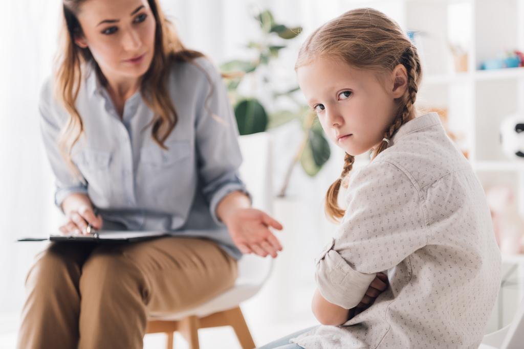 psychologist talking to angry child with crossed arms while she looking at camera - Photo, Image