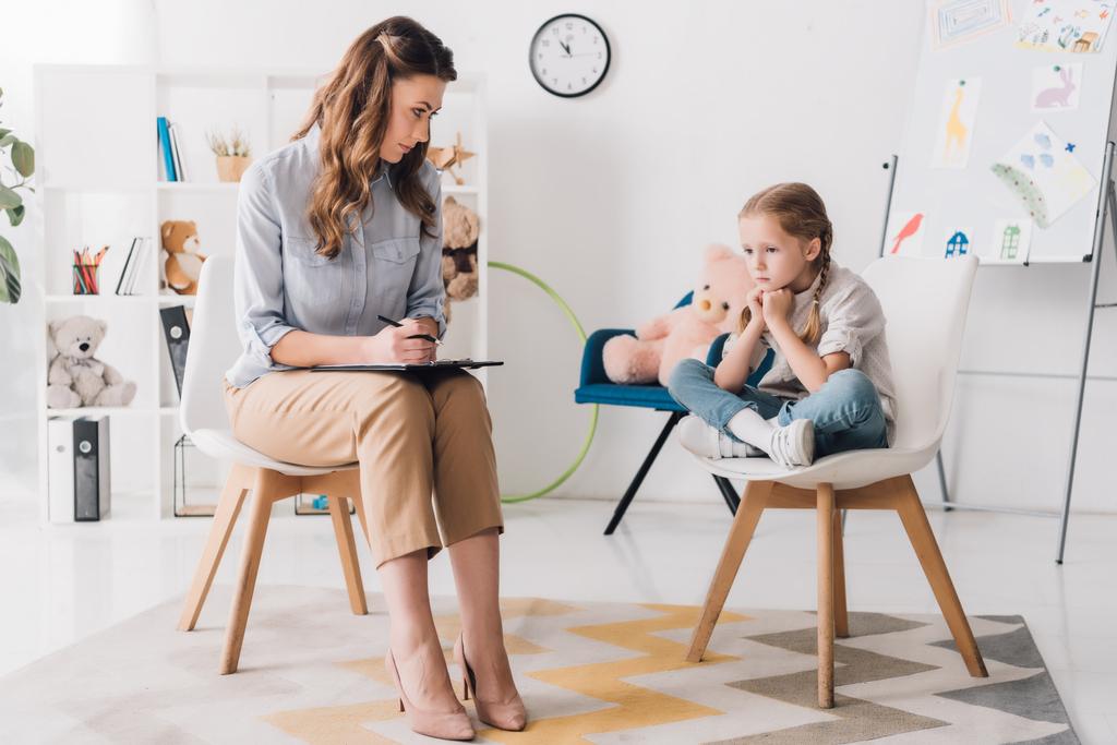 ernsthafter Erwachsenenpsychologe mit Klemmbrett sitzt neben kleinem depressiven Kind im Büro - Foto, Bild