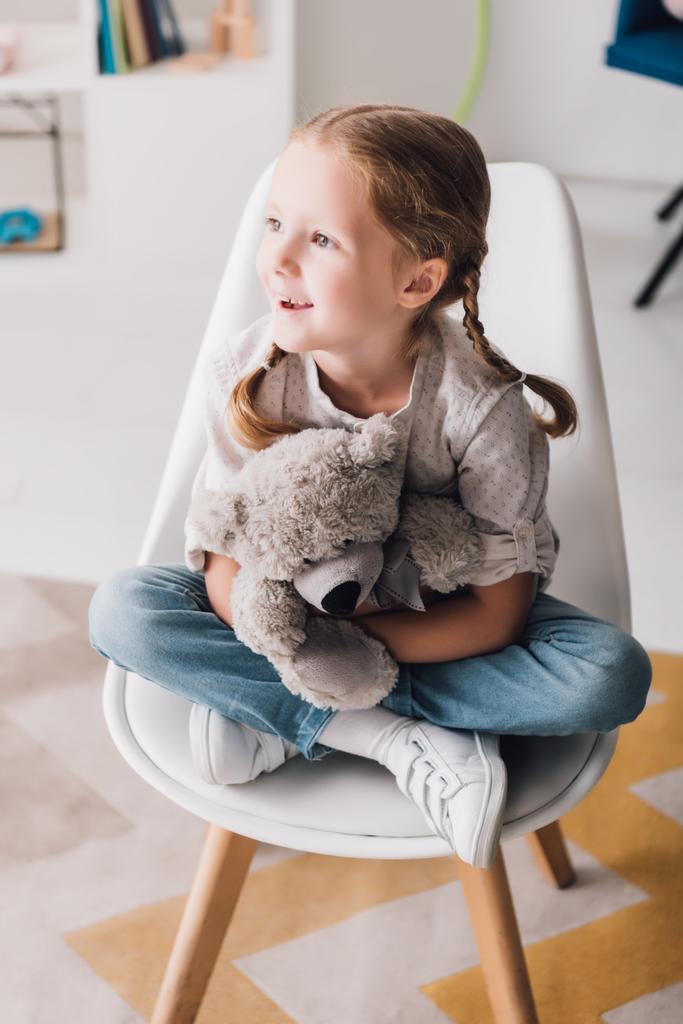 boldog kis gyerek ül a székre, és átfogó ő medve plüss - Fotó, kép