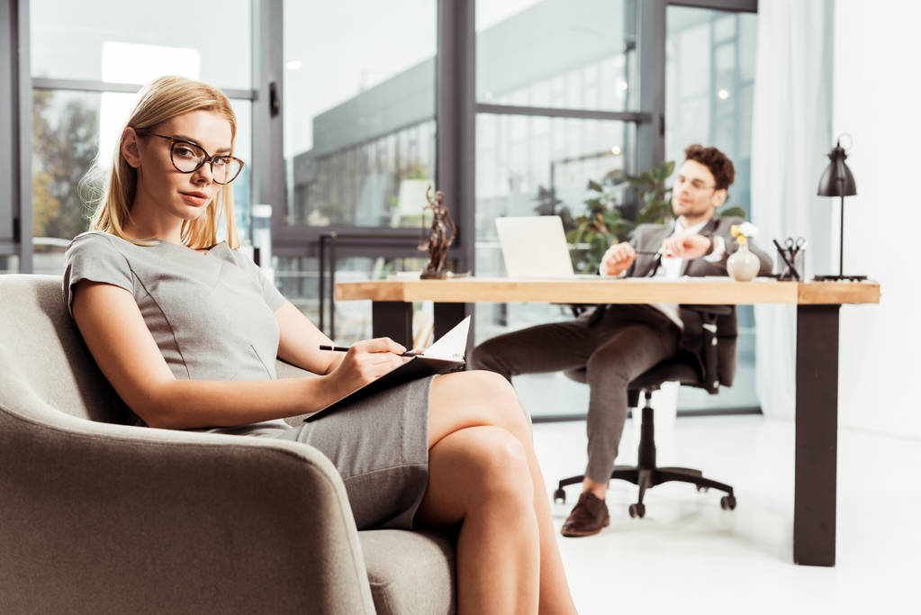 businesswoman with notebook looking at camera while sitting in armchair with colleague at workplace in office - Photo, Image