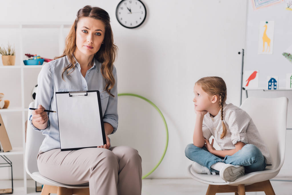 Psychologe hält leeres Klemmbrett, während er neben depressivem Kind im Büro sitzt - Foto, Bild