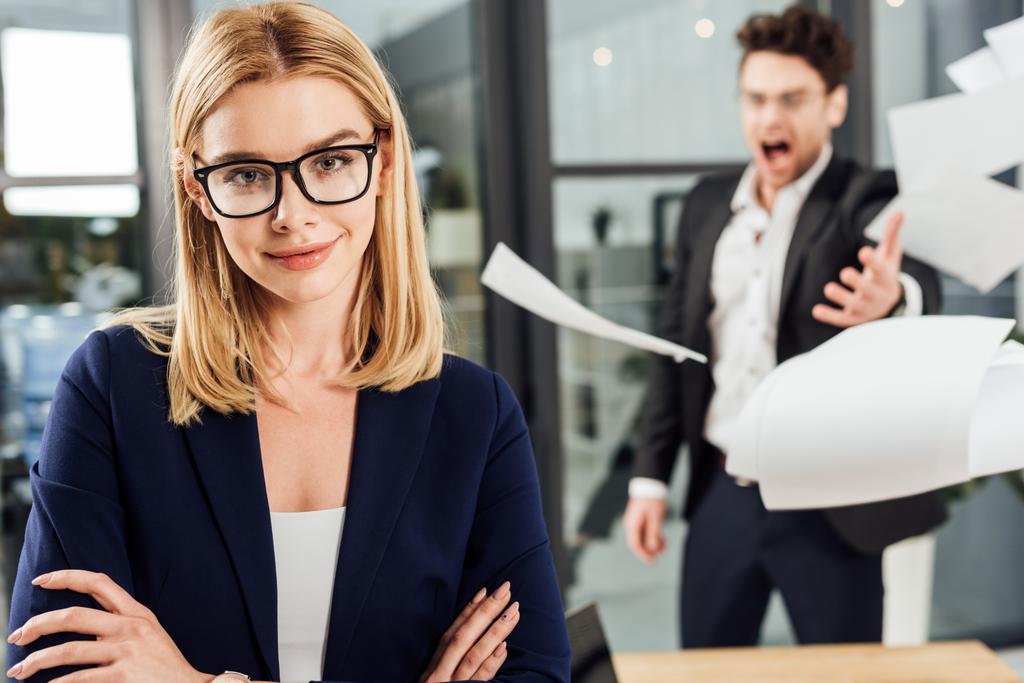избирательный фокус улыбающейся бизнесвумен в очках и злой кричащий босс бросает бумаги позади в офисе
 - Фото, изображение