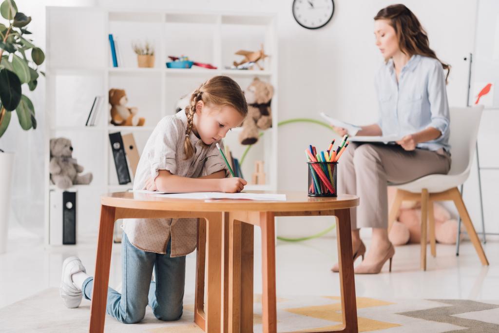 little child drawing while psychologist with clipboard sitting blurred on background - Photo, Image