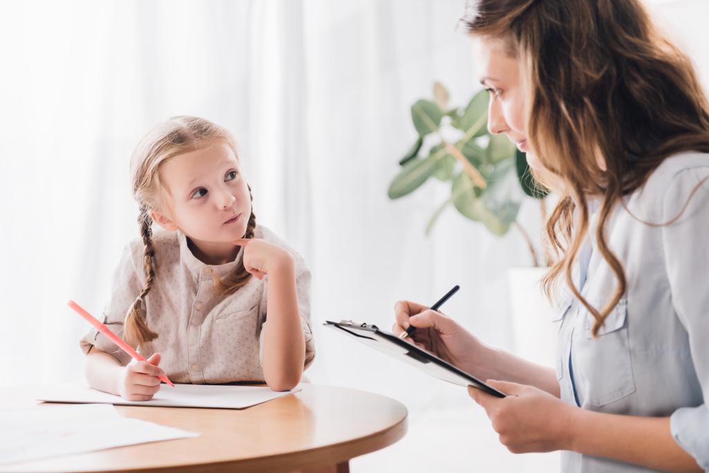 psicólogo com área de transferência sentado perto de criança e conversando enquanto ela desenha com lápis de cor
 - Foto, Imagem
