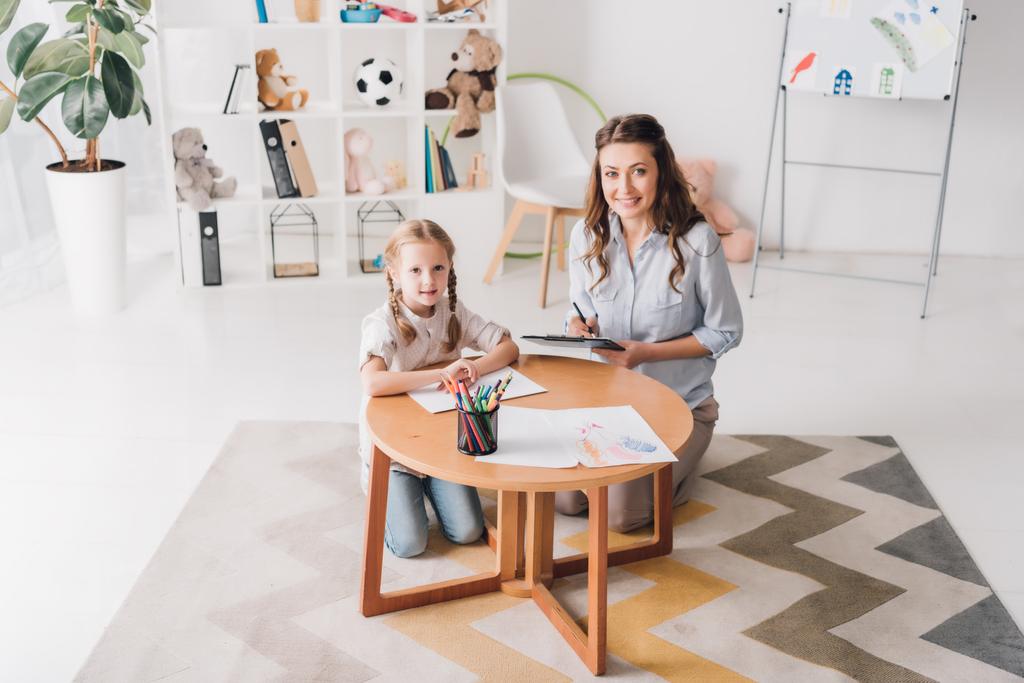 happy psychologist with clipboard sitting near little child while she drawing with color pencils and looking at camera - Photo, Image