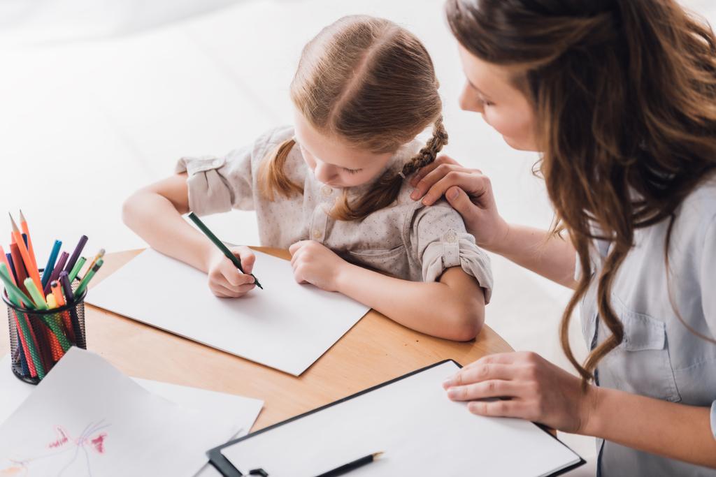 vista ad alto angolo di psicologo con appunti seduto vicino al bambino mentre lei disegno con matite di colore
 - Foto, immagini