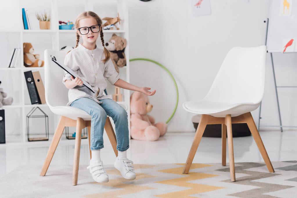 adorable little child in eyeglasses with clipboard sitting on chair and looking at camera - Photo, Image