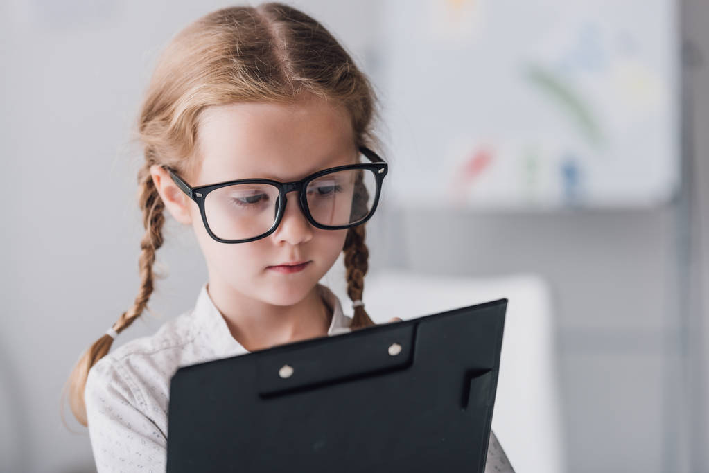close-up portrait of serious little child in eyeglasses writing in clipboard - Photo, Image