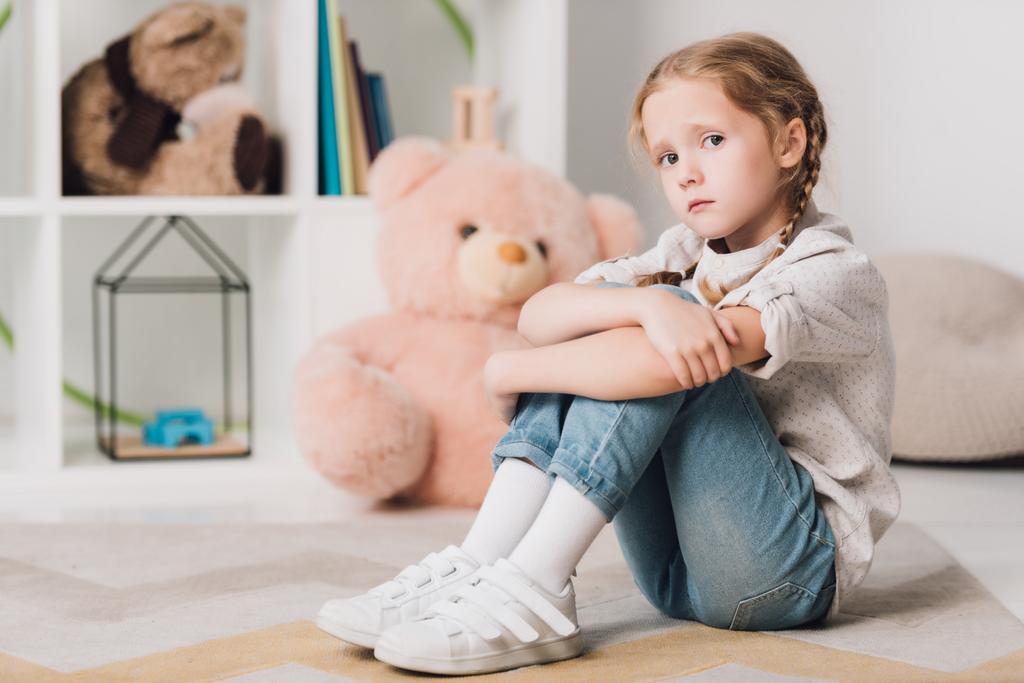 lonely little child sitting on floor in front of shelves with toys and looking at camera - Photo, Image