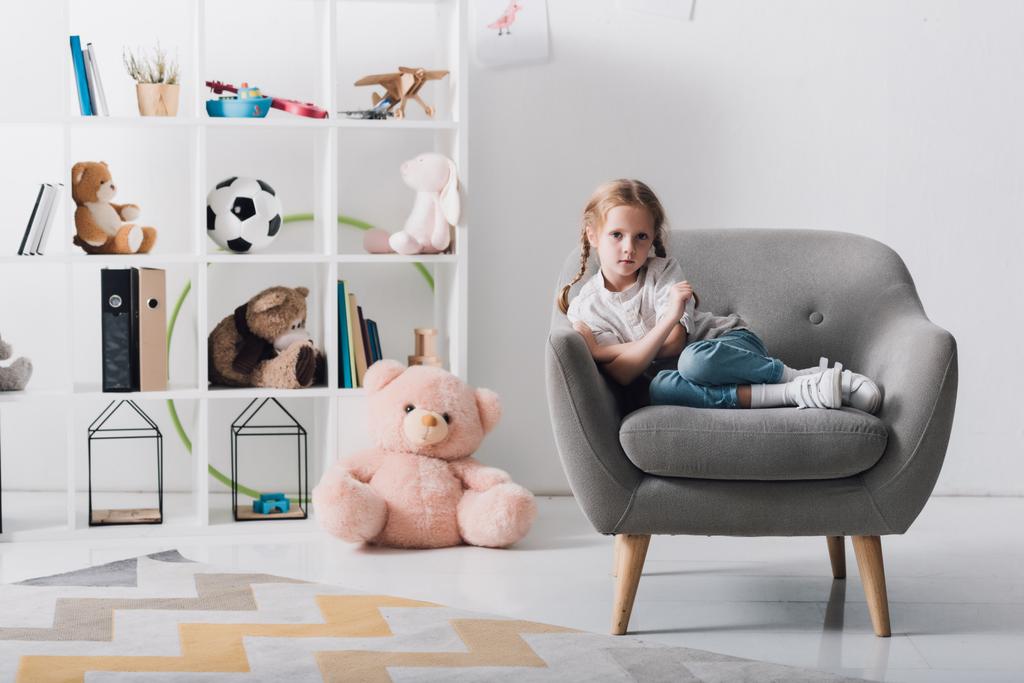 samotny małe dziecko siedząc na fotelu przed półkami z zabawkami i patrząc na kamery - Zdjęcie, obraz