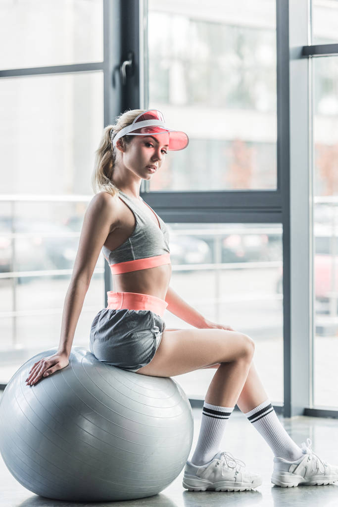 Sportvrouw in vizier hoed zittend op grijs fitness bal op sportschool - Foto, afbeelding