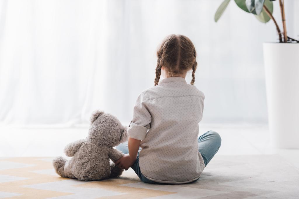 вид сзади на маленького ребенка, сидящего на полу со своей игрушкой плюшевого мишки
 - Фото, изображение