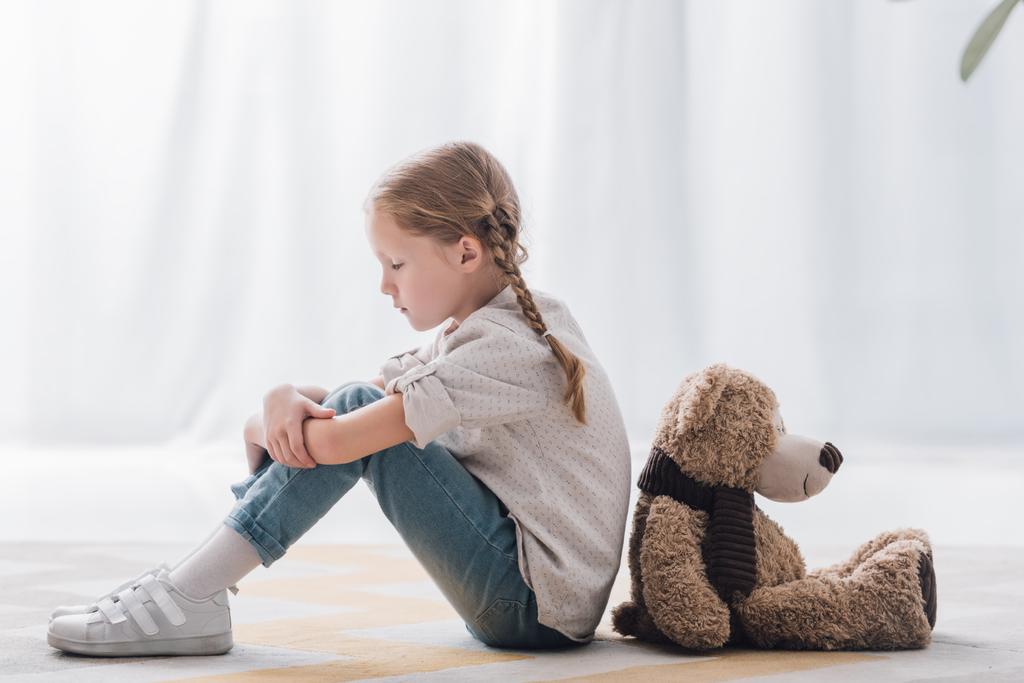 Seitenansicht eines traurigen kleinen Kindes, das Rücken an Rücken mit Teddybär auf dem Boden sitzt - Foto, Bild