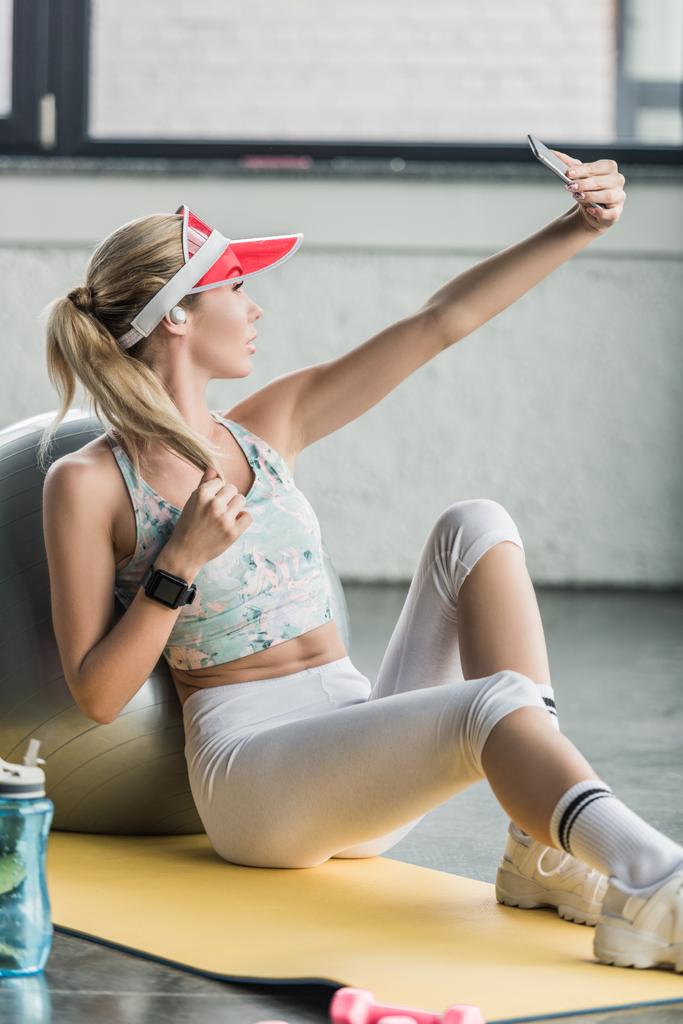 Seitenansicht einer Sportlerin mit Smartwatch, die in der Nähe eines Fitness-Balls im Fitnessstudio ein Selfie mit dem Smartphone macht - Foto, Bild