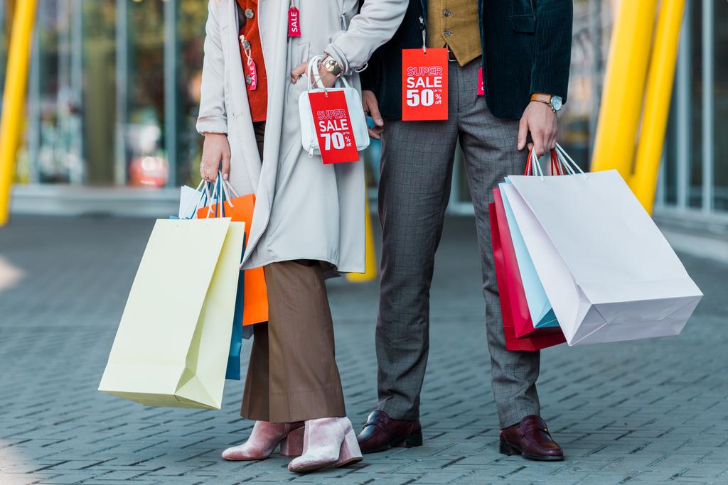 bijgesneden weergave van stijlvolle elegante paar met boodschappentassen en verkoop tags poseren in de buurt van shopping center - Foto, afbeelding