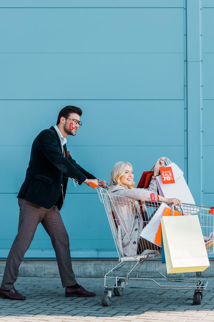 belo casal elegante com sacos de compras e etiquetas de venda se divertindo com carrinho de compras na parede azul
 - Foto, Imagem