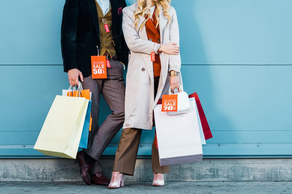 vista recortada de pareja elegante sosteniendo bolsas de compras con etiquetas de venta y posando en la pared azul
 - Foto, imagen