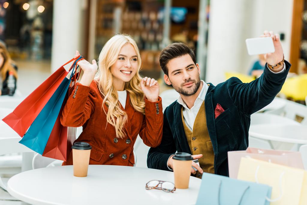 modieuze paar met boodschappentassen en koffie te gaan nemen selfie op smartphone in winkelcentrum - Foto, afbeelding