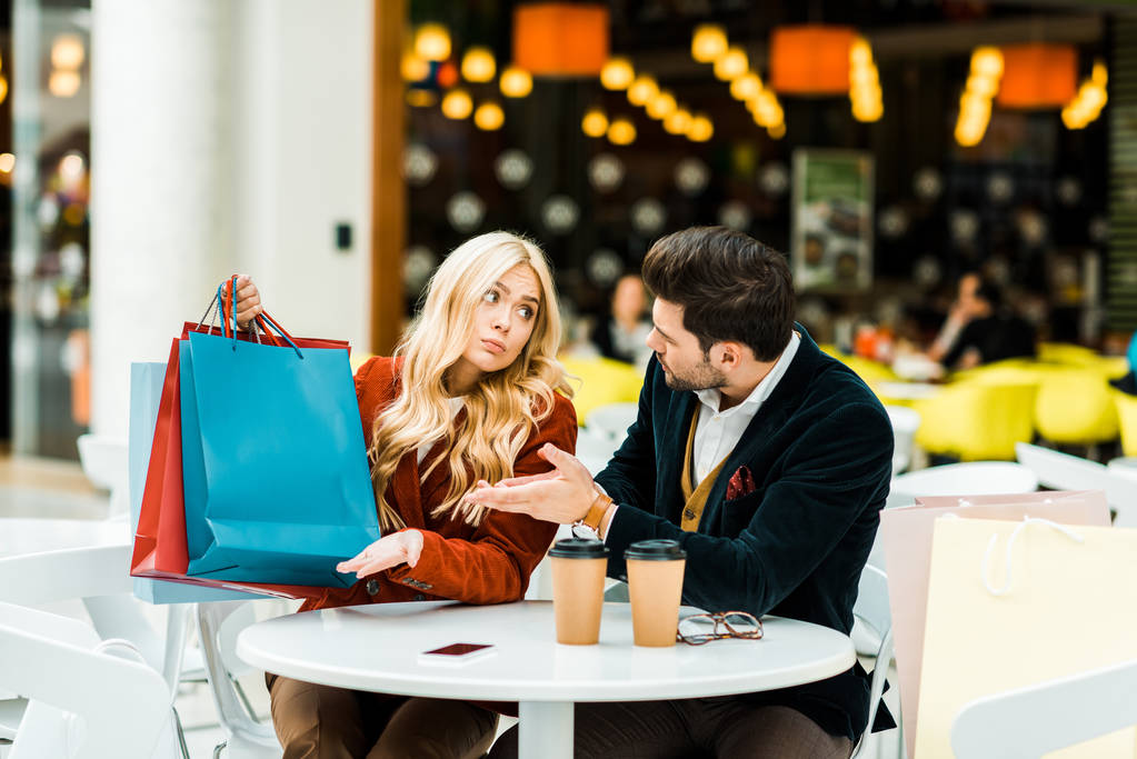 mulher confusa mostrando sacos de compras para o homem no café no shopping
 - Foto, Imagem