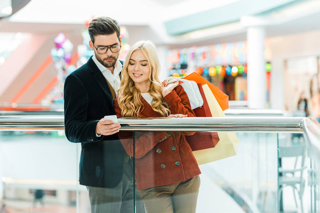 jeune couple élégant avec sacs à provisions en utilisant smartphone dans le centre commercial
 - Photo, image