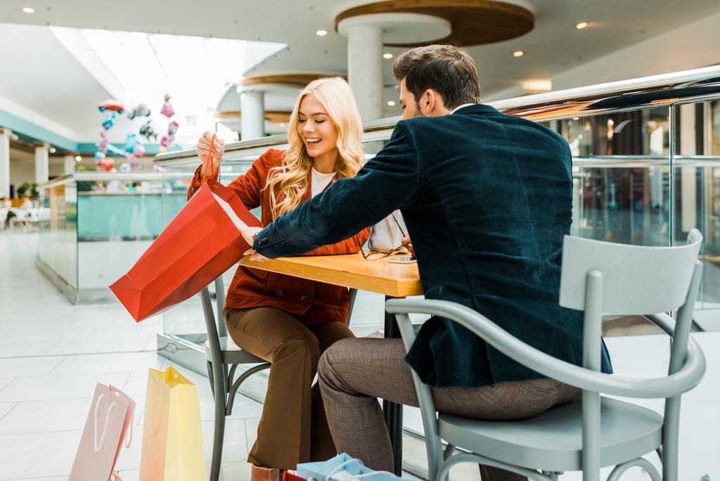 belle femme heureuse montrant quelque chose dans le sac à provisions pour petit ami tout en étant assis dans le café dans le centre commercial
 - Photo, image