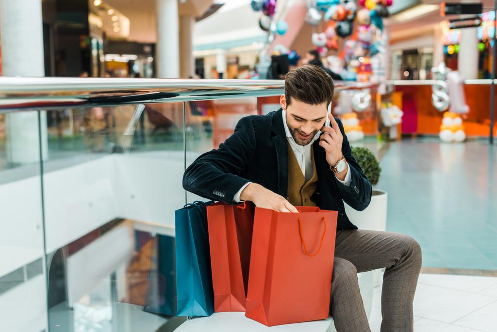 handsome man talking on smartphone and looking into bags while sitting in shopping mall  - Photo, Image
